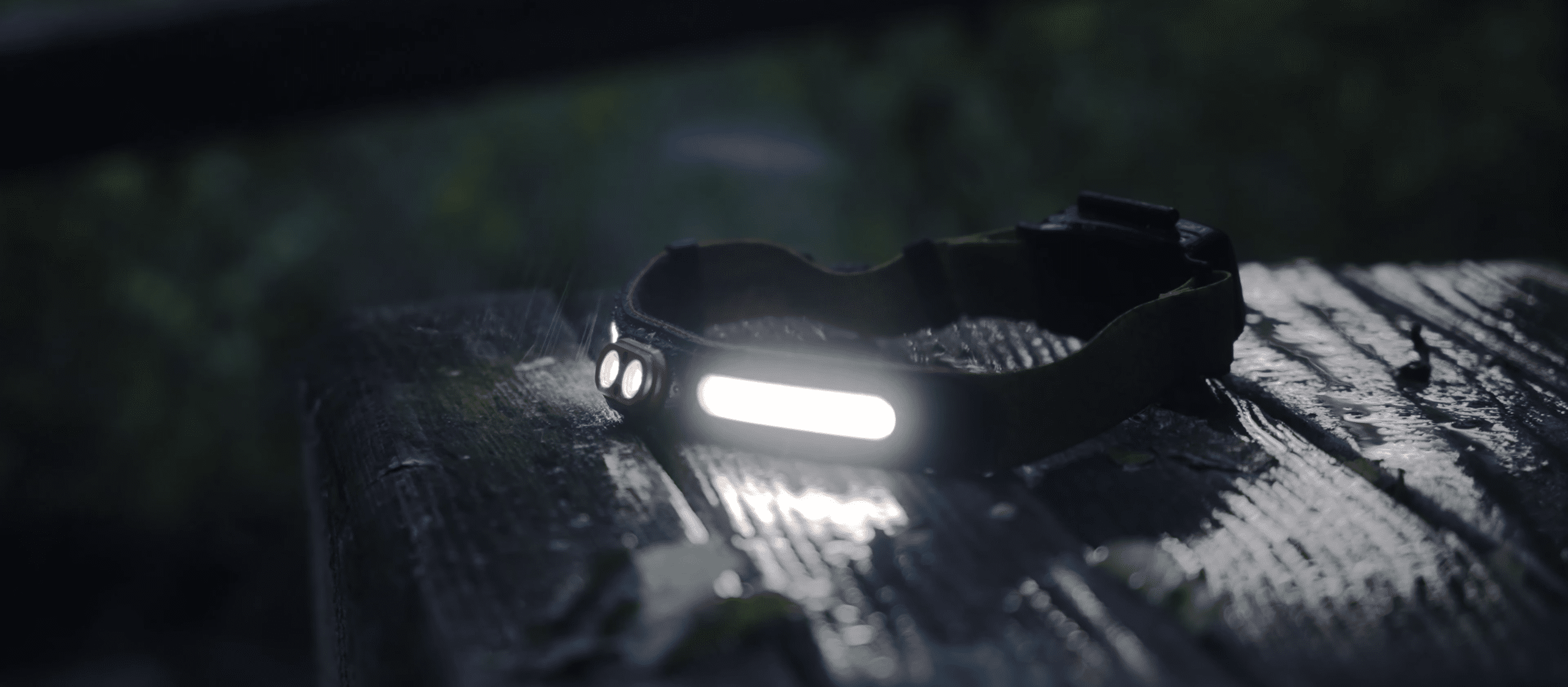 A headlamp with a bright LED light rests on a wet wooden surface outdoors, featured in Overland News, with blurred greenery in the background.