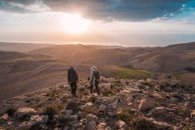 Two hikers with backpacks walk along a rocky, sunlit hillside, overlooking rolling, barren mountains at sunset. The vast landscape stretches toward the horizon—an inspiring scene fit for an Overland News adventure.