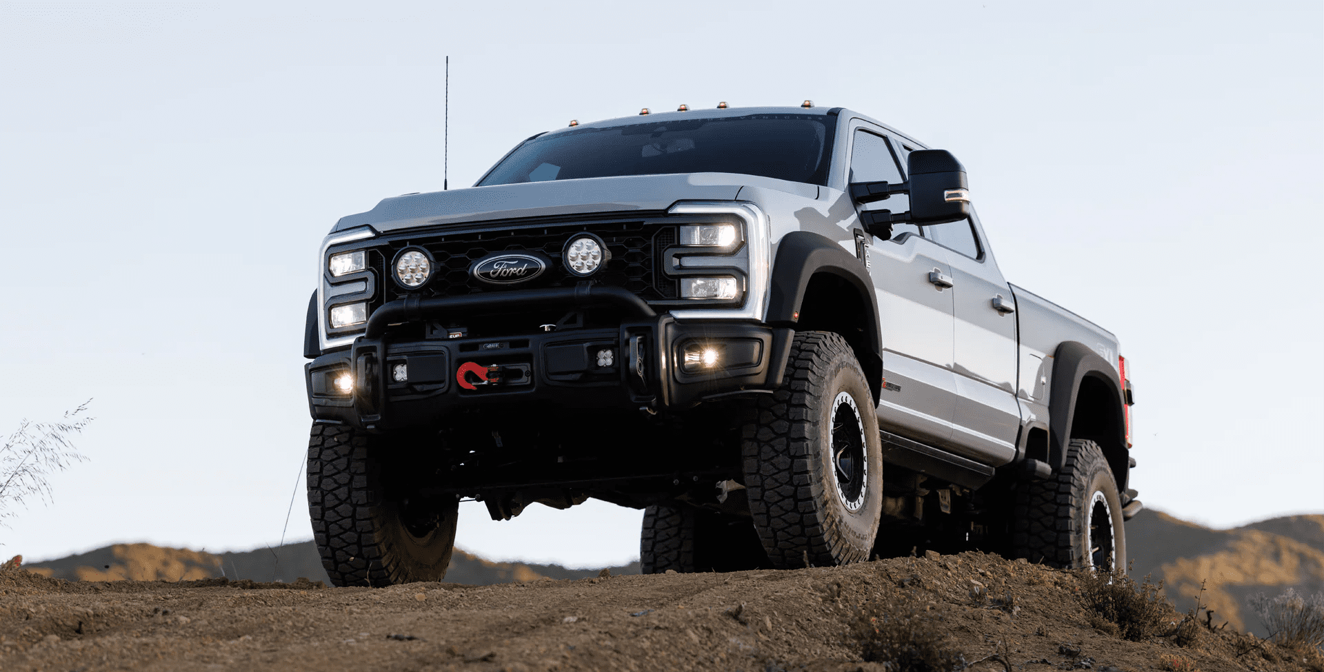 A silver Ford pickup truck with off-road tires and extra lights, featured in Overland News, is parked on a dirt hill and viewed from a low angle against a clear sky.