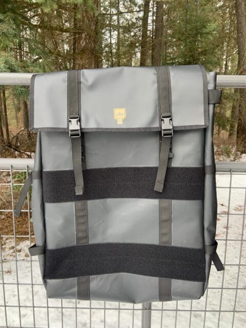 A large gray backpack with black straps and buckles hangs on a metal fence outdoors. Snow covers the ground as tall evergreen trees stand in the background, creating a scene worthy of Overland News.