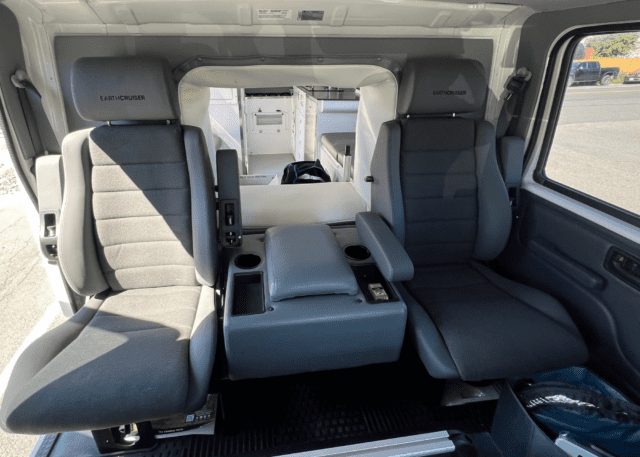 Two gray, padded car seats with armrests and EarthCruiser branding are positioned inside a vehicle, separated by a central console with cup holders and storage. Sunlight streams in through the nearby window, highlighting this Overland News feature.