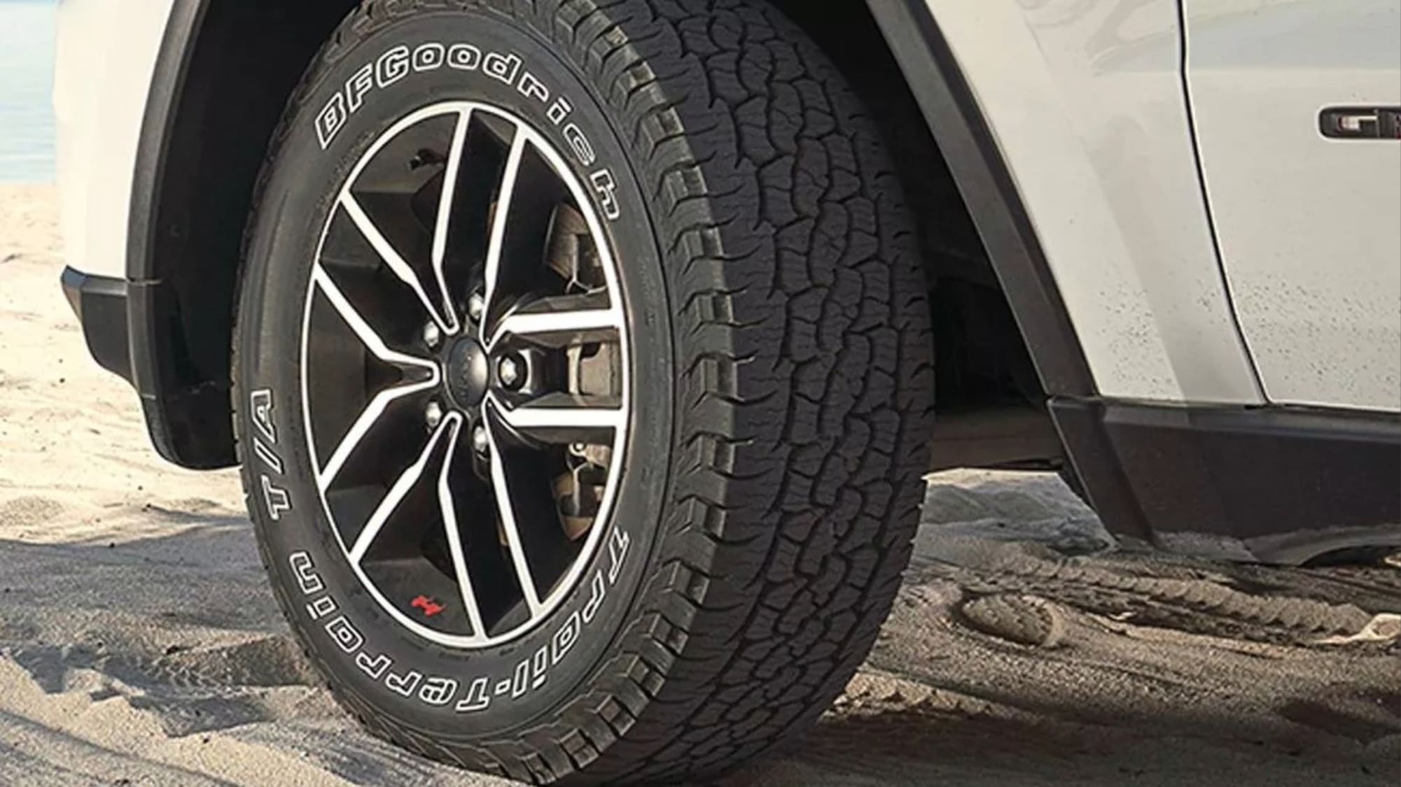 Close-up of a black BFGoodrich All-Terrain T/A tire mounted on a black and silver alloy wheel of an SUV, parked on sandy ground near water—perfect for your next Overland News adventure.