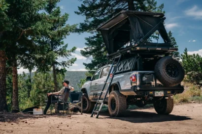 A person sits in a camp chair beside a campfire near a silver pickup truck with a rooftop tent, featured in Overland News, parked among pine trees in a scenic outdoor setting. A ladder leads to the tent and camping gear is nearby.