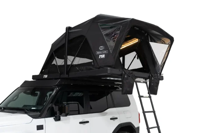 A black rooftop tent is mounted on top of a white SUV, with a ladder attached for access. Featured in Overland News, the tent has clear windows and is partially open, showing its structure and providing space for camping.