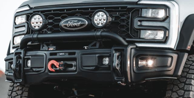 Close-up of a black Ford truck front bumper with off-road features, including a winch, red hook, LED lights, and a bold grille displaying the Ford logo—perfect for Overland News coverage. Tires and headlight are partially visible.