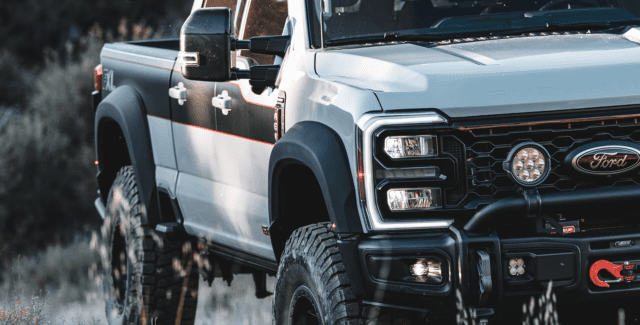 A close-up of the front and side of a lifted white Ford pickup truck with black fender flares, off-road tires, and custom lights, featured by Overland News, parked outdoors in a grassy area.