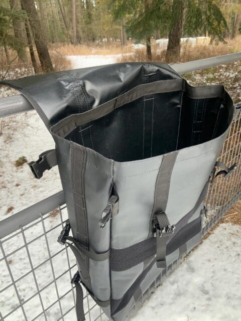 A large, empty, black waterproof bag with straps hangs over a metal fence in a snowy forested area. Pine trees and patches of snow are visible in the background, creating the perfect backdrop for an Overland News photo.