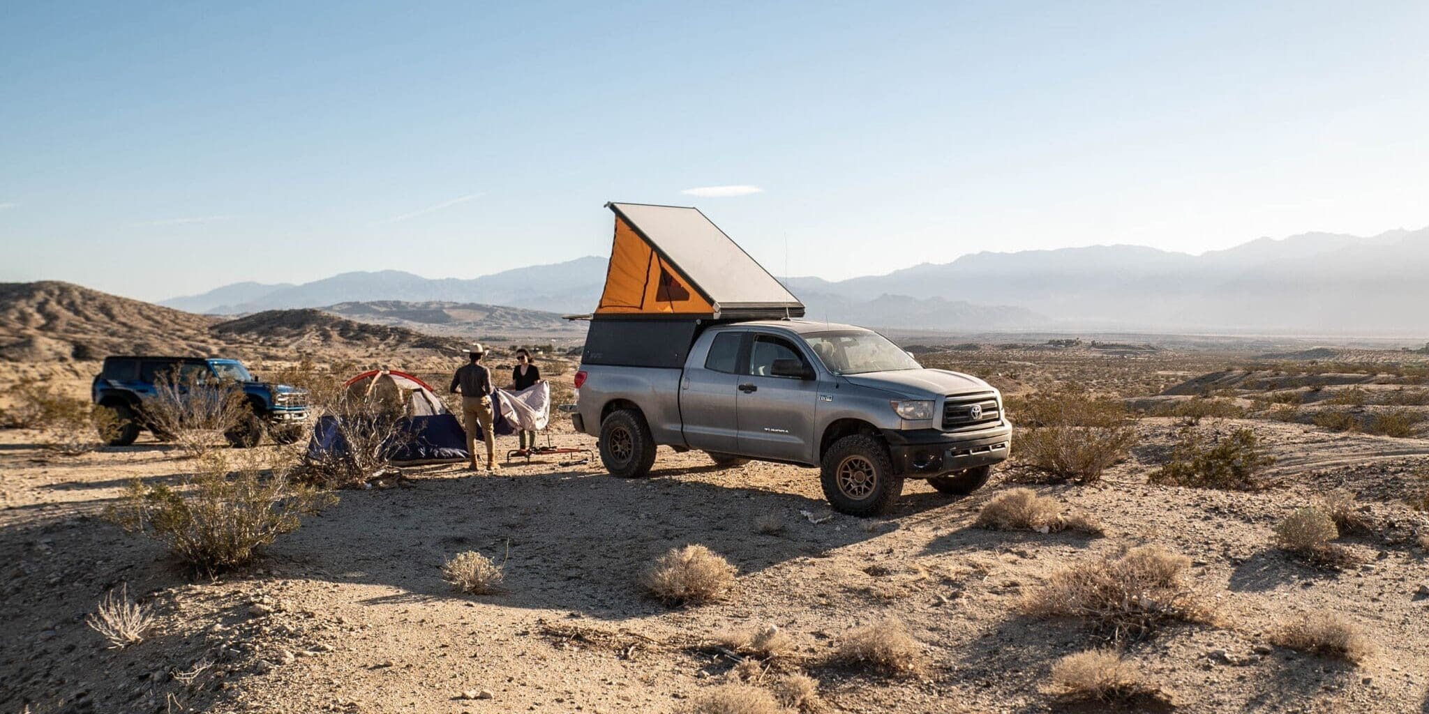 A silver pickup truck with a rooftop tent is parked on a desert landscape. Nearby, people stand by a blue tent with mountains in the background under a clear sky—a classic scene for Overland News enthusiasts.