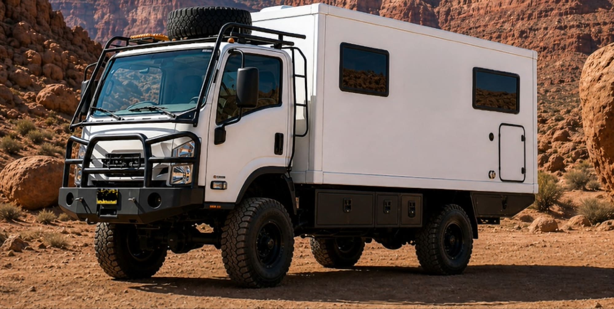 A white off-road expedition truck with large tires, a roof rack, and a spare tire is parked on a dirt path in rocky desert terrain. Featured in Overland News, the boxy camper module has tinted windows for privacy and rugged style.