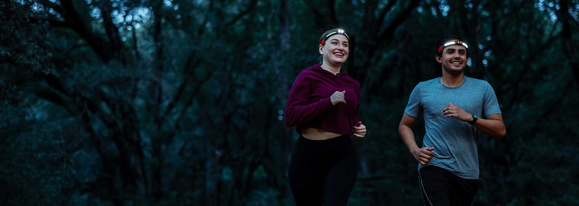 Two people wearing headlamps and athletic clothes run through a dark, wooded area, smiling as they exercise together in the early morning or evening—a scene worthy of Overland News.