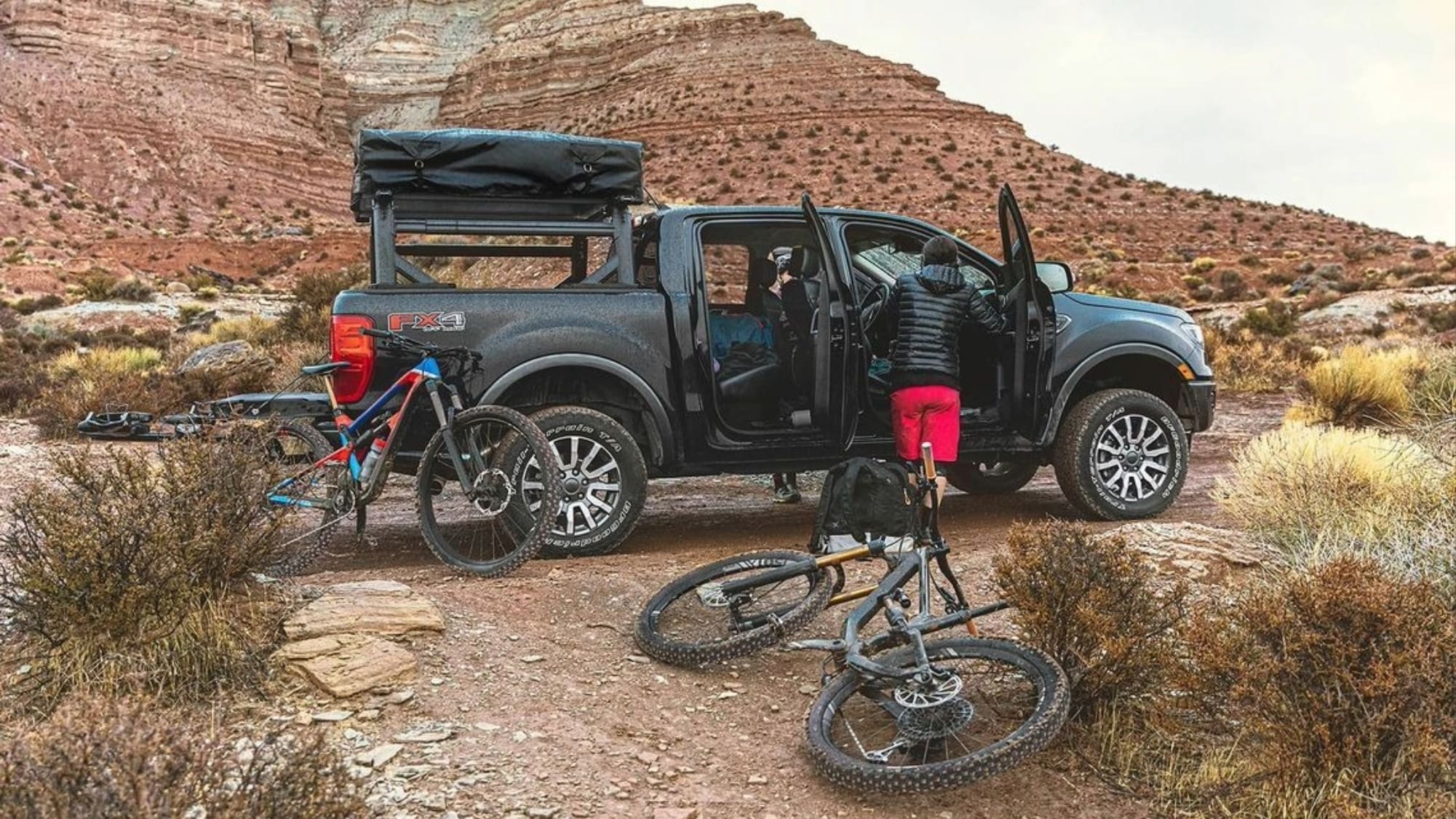 A pickup truck is parked in a rocky desert landscape with two mountain bikes nearby. One person in red shorts stands by the open door, ready for an Overland News-worthy adventure. Rugged hills and desert plants fill the background.
