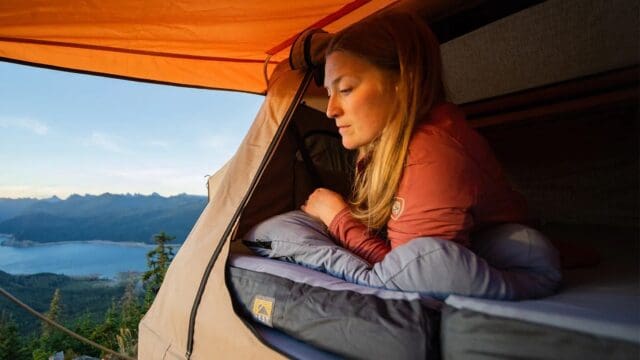 A woman lies inside a tent, resting on her elbow and looking out over a scenic mountain landscape with a lake at sunset. She appears relaxed, surrounded by camping gear—an ideal moment for any Overland adventure enthusiast.
