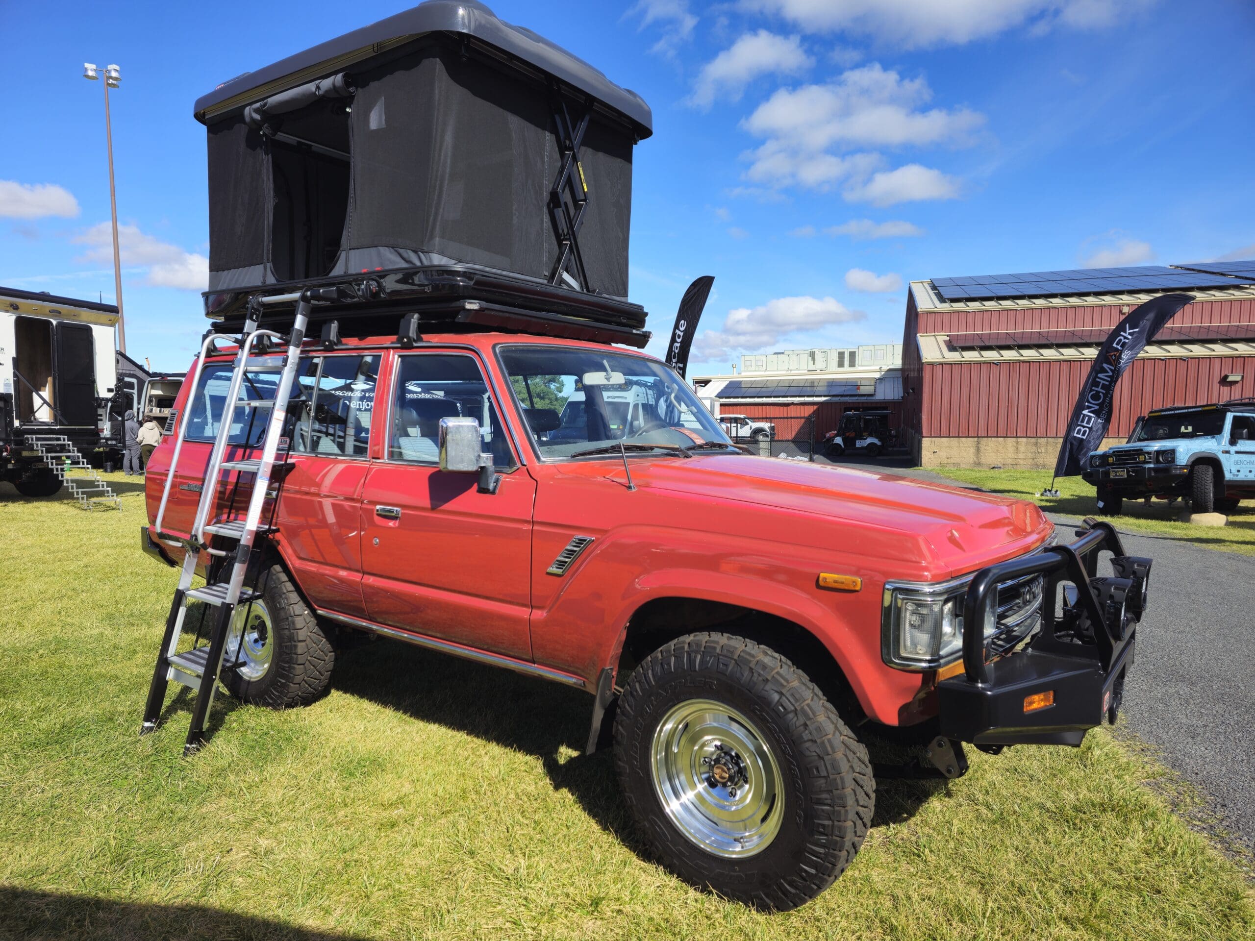 A red off-road vehicle with a rooftop tent and ladder is parked on grass beside a building at Overland Expo. The tent is open, revealing a black enclosure above the car. Other vehicles and banners are visible in the background on this sunny day.