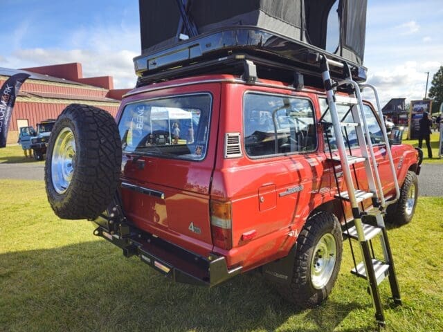 A red off-road vehicle with a roof tent and ladder set up at Overland Expo. A spare tire is mounted on the back, with other vehicles and buildings visible in the background.