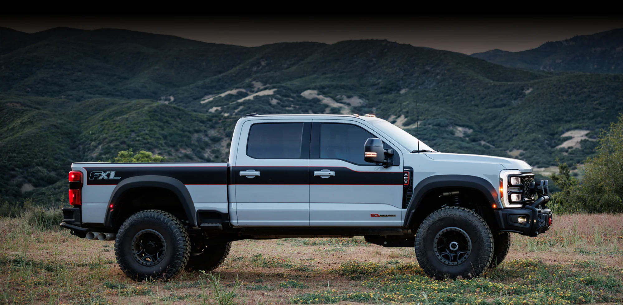 A white Super Duty pickup truck with black accents and large off-road tires is parked on grassy terrain, with green mountains in the background—a rugged scene inspired by AEV FXL design.
