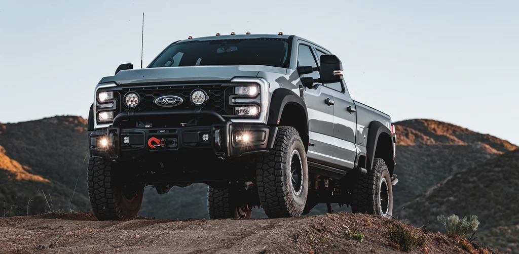 A lifted gray AEV FXL Super Duty Ford pickup truck with off-road tires and additional front lights is parked on a dirt trail, with hills and a clear sky in the background.
