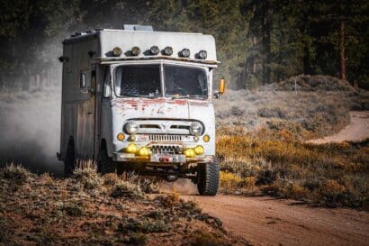 A rugged, vintage 1953 Ford off-road truck with extra lights and large tires drives on a dusty dirt road through a dry, grassy landscape with trees in the background.