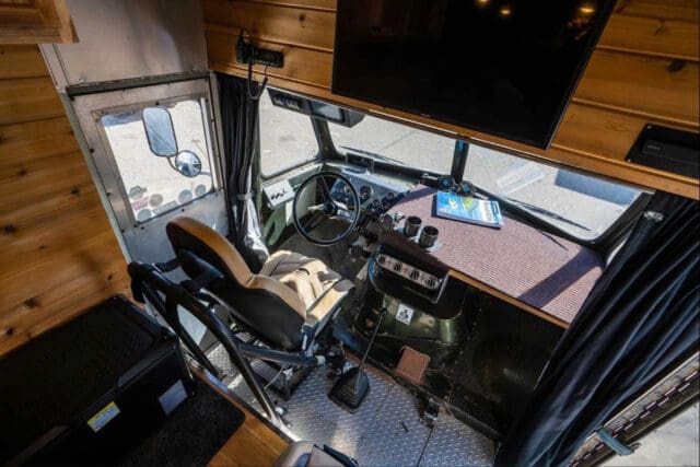 View of a retro-style RV driver’s cockpit with a vintage steering wheel, cushioned seat, manual gear shift, wood-paneled walls, and a large TV above—classic Chuckles Garage vibes. A magazine and phone rest on the dashboard.