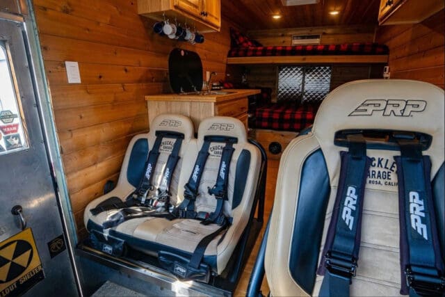 Interior of a custom van by Chuckles Garage, featuring wood paneling, two beige PRP bucket seats with harnesses, a compact kitchen area, shelves with mugs, and a cozy bed in the back topped with a red-and-black plaid blanket.