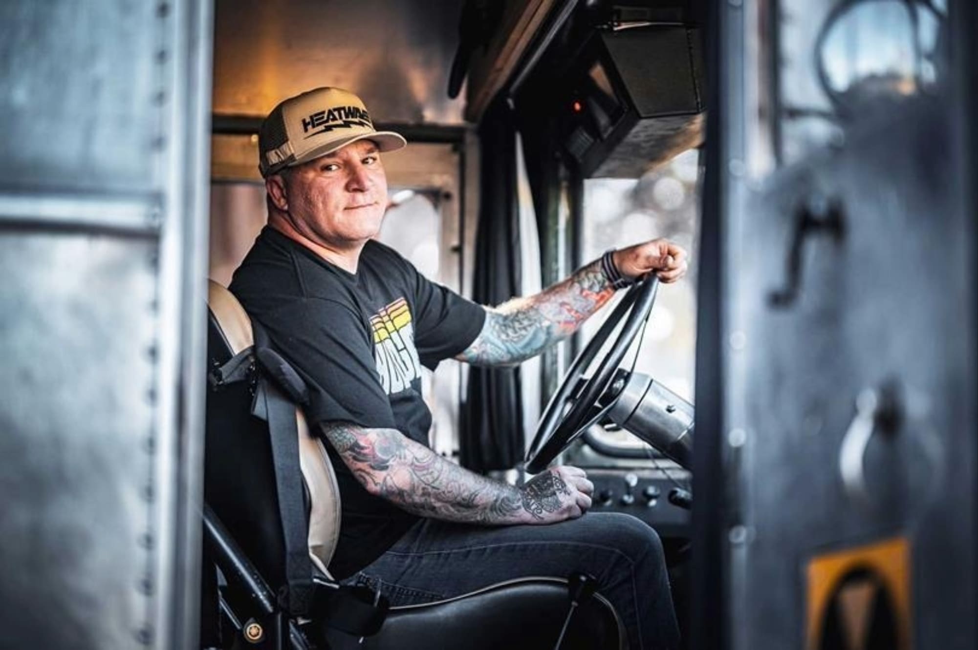 A man with tattoos, wearing a cap and a black Chuckles Garage t-shirt, sits in the driver’s seat of a vehicle, holding the steering wheel and looking towards the camera. The interior of the vehicle is visible around him.