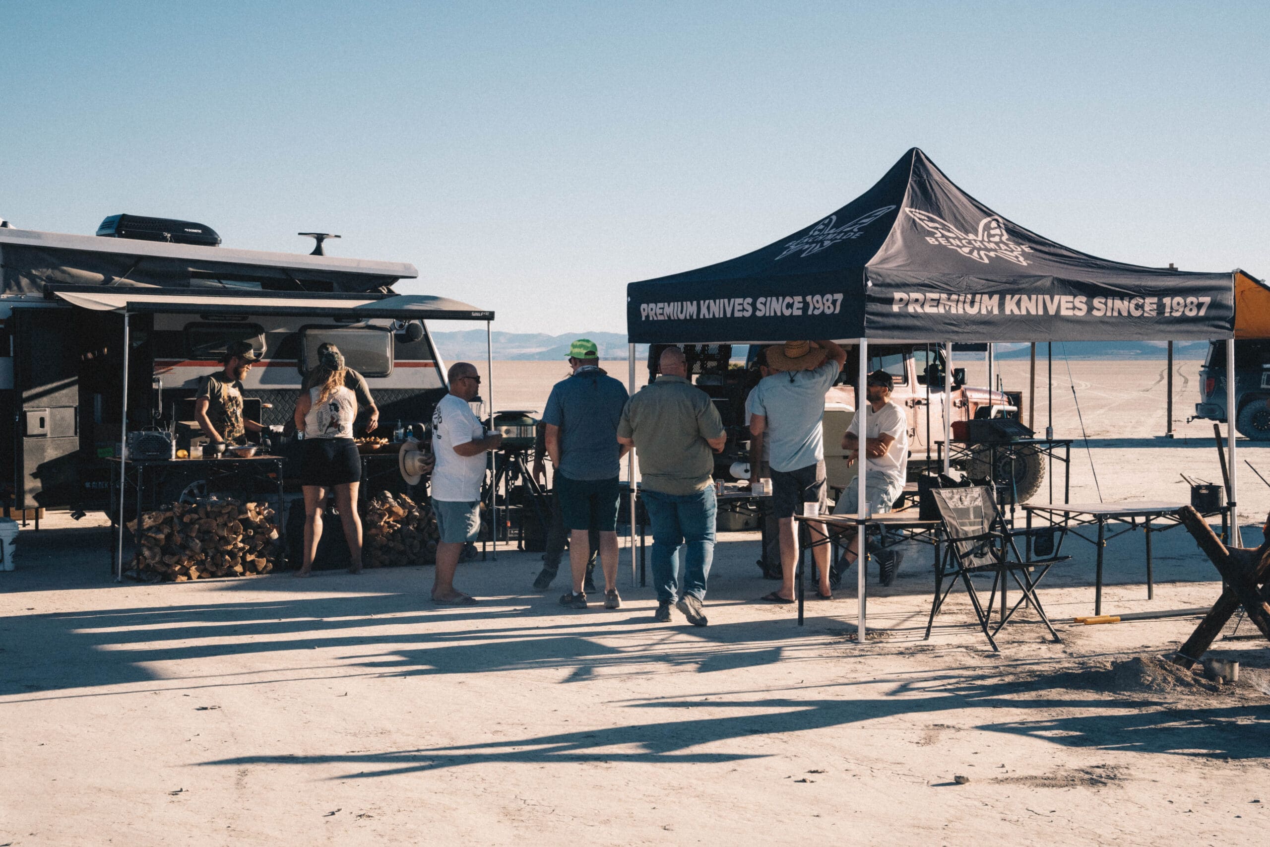 A group of people gather under a tent labeled Premium Knives Since 1987 near an outdoor kitchen setup and camper, enjoying a Trail Feast on the sunlit expanse of Alvord Playa with mountains in the background. The scene reflects the spirit of adventure and quality craftsmanship found in every Benchmade blade.