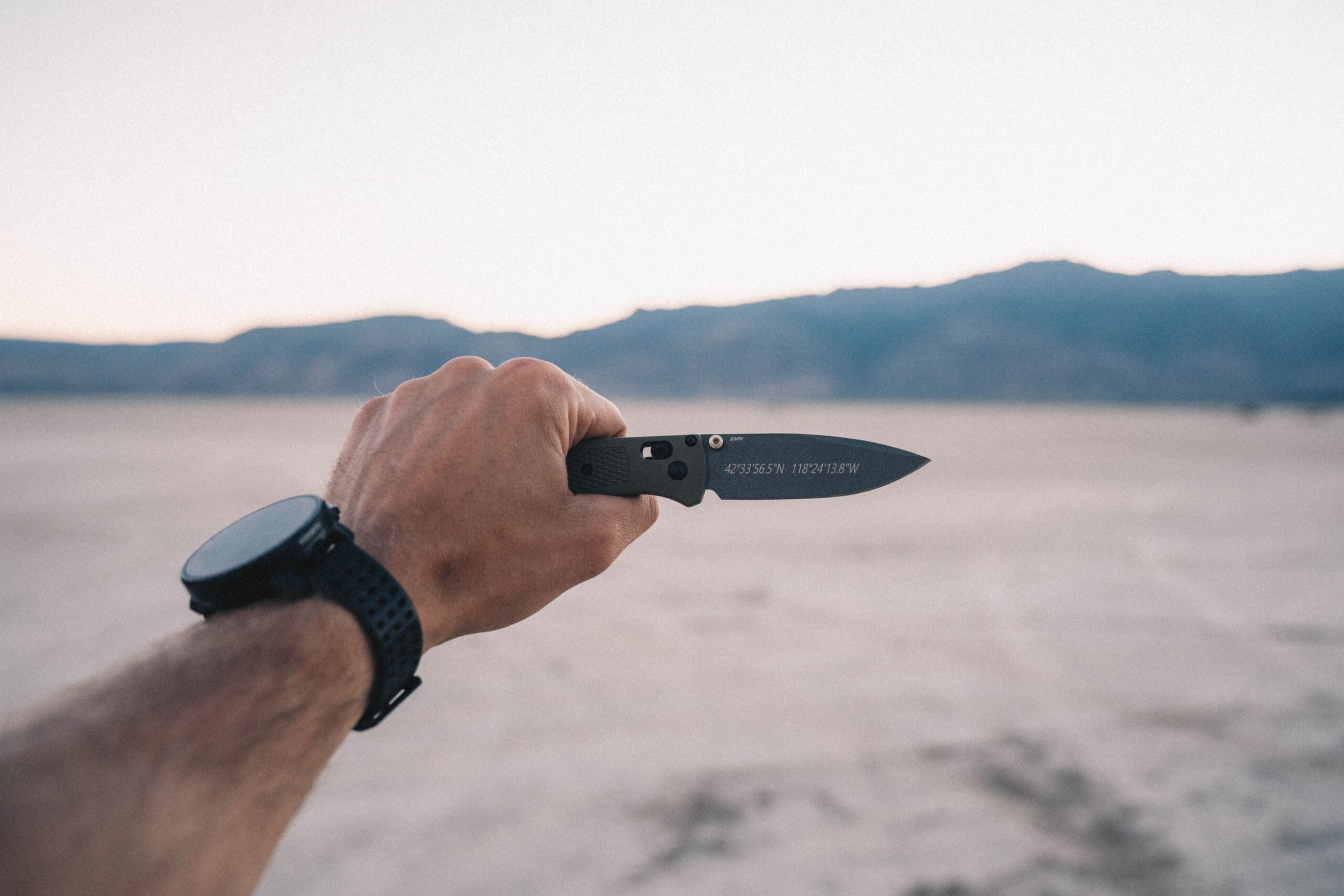 A person wearing a black watch holds a Benchmade folding knife outdoors on the Alvord Playa, with a dry, open landscape and distant mountains under a pale sky in the background.