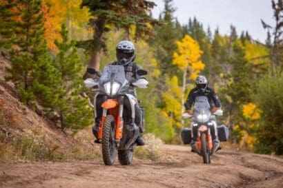Two people ride adventure motorcycles on a dirt trail through a forest with green and yellow trees, reminiscent of routes featured in overland events and rallies. Both riders wear helmets and protective gear as they navigate the rugged, natural trail surrounded by vibrant autumn foliage.