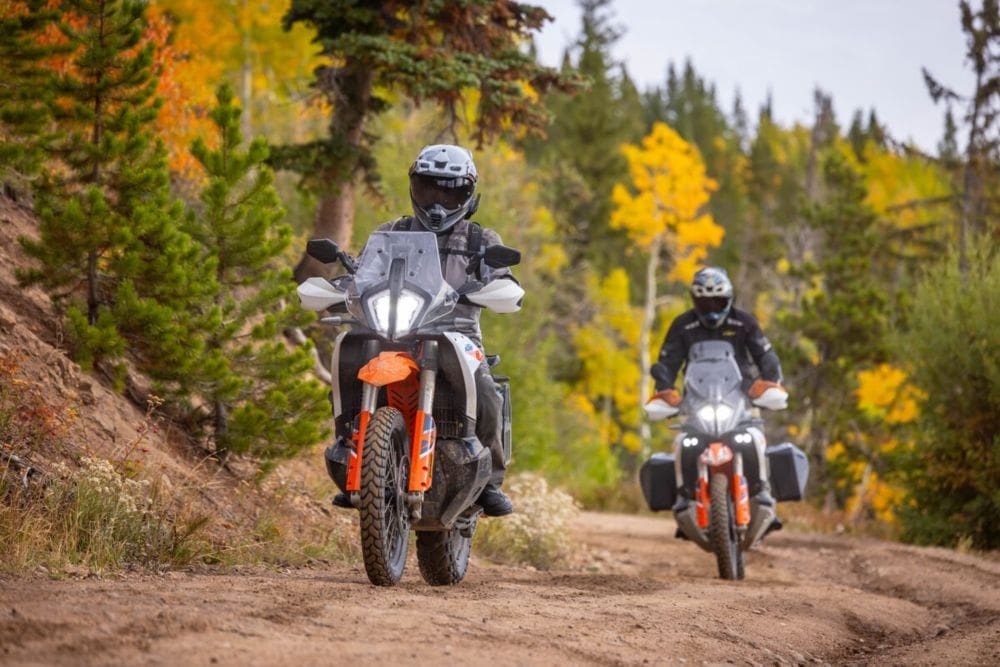 Two people ride adventure motorcycles on a dirt trail through a forest with green and yellow trees, reminiscent of routes featured in overland events and rallies. Both riders wear helmets and protective gear as they navigate the rugged, natural trail surrounded by vibrant autumn foliage.