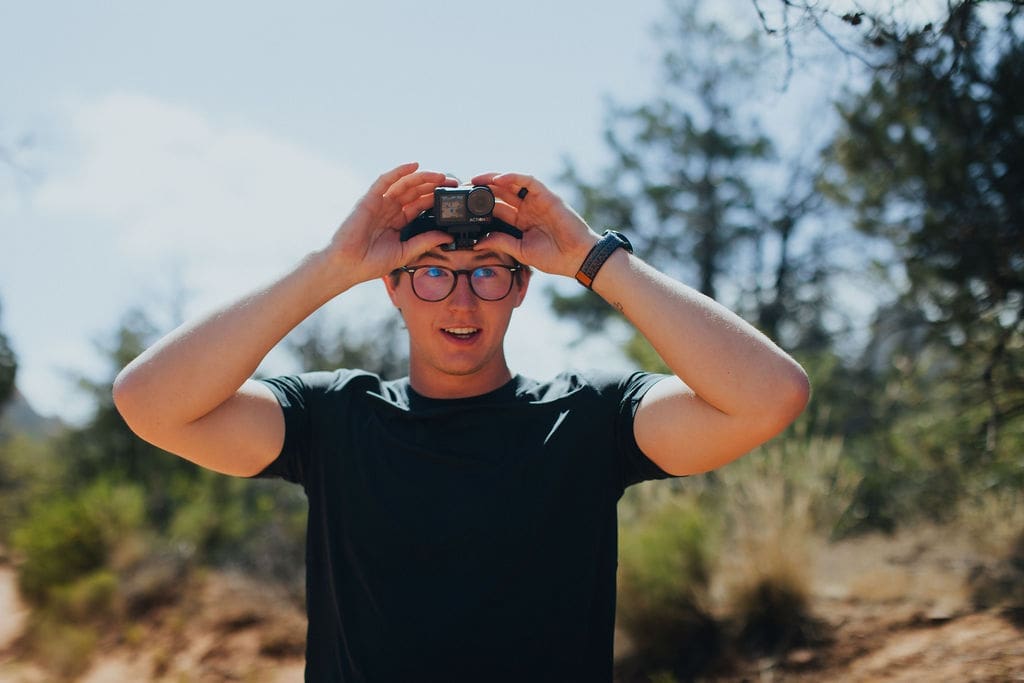 A person wearing glasses and a dark shirt smiles while holding a small camera above their head outdoors, capturing the spirit of 36-Hour Adventures amid blurred trees and dry grass in the background.