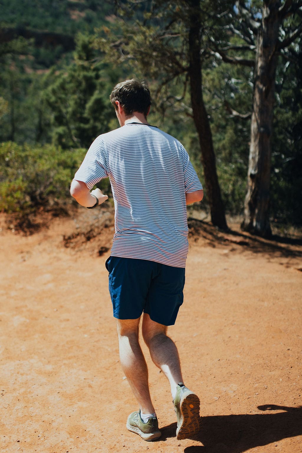 A person wearing a striped shirt and blue shorts walks or jogs on a sunny dirt trail surrounded by trees and greenery, embracing the spirit of 36-Hour Adventures.
