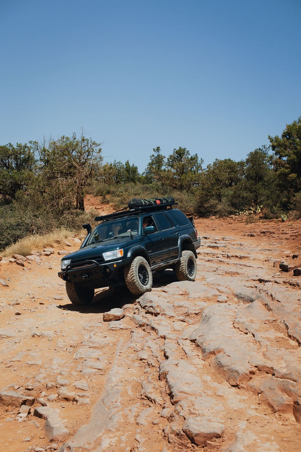 A black SUV with off-road tires embarks on one of its 36-Hour Adventures, driving over rocky, uneven terrain in a desert landscape with sparse trees and a clear blue sky.