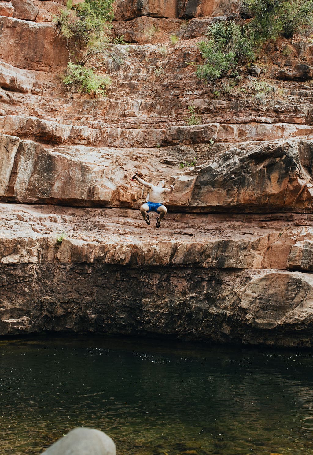 A person in a white shirt and blue shorts jumps off a rocky cliff toward a body of water below, surrounded by rugged, layered rock formations and sparse greenery—capturing the thrill of 36-Hour Adventures in the heart of nature.