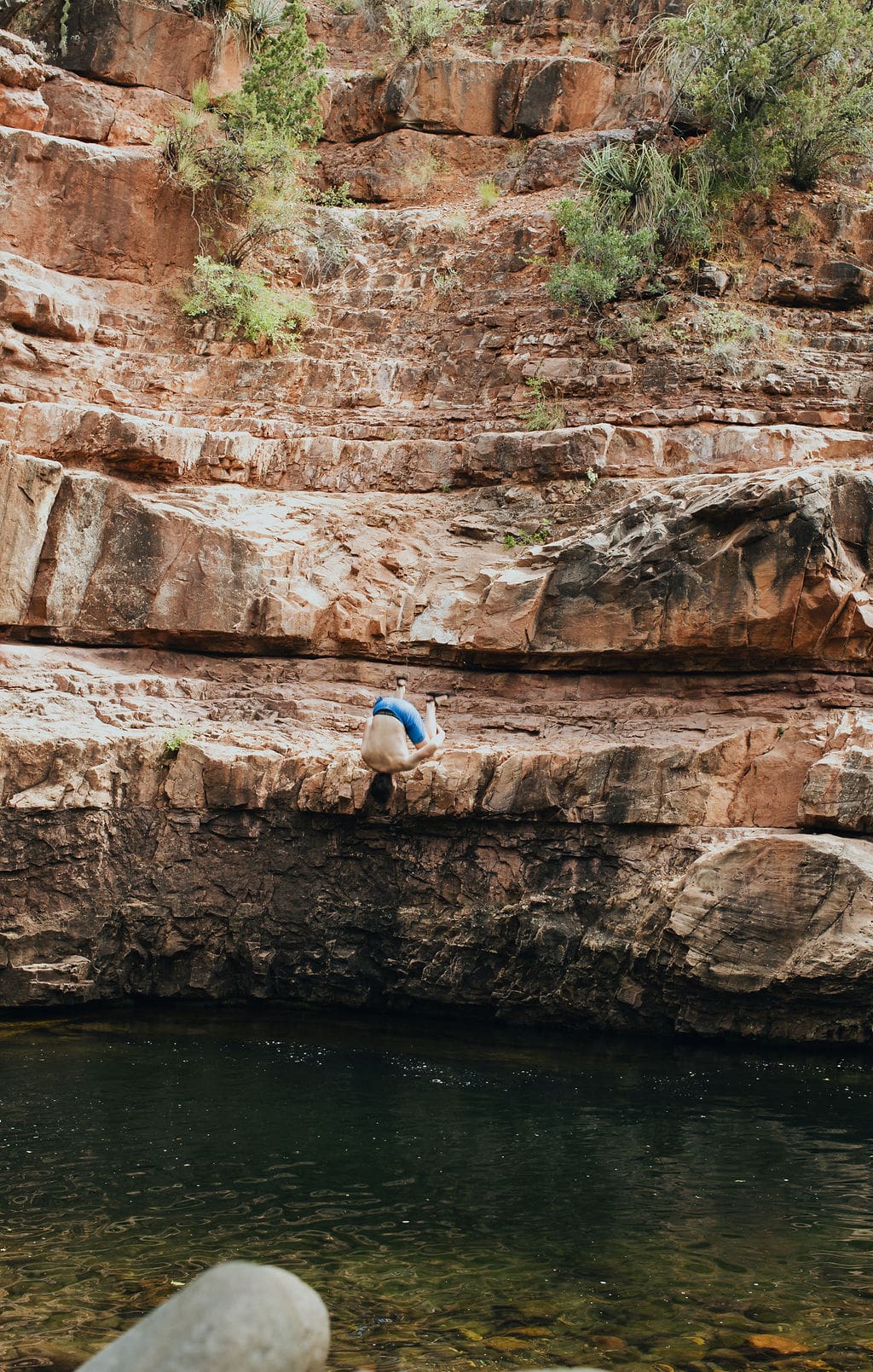A person in blue shorts crouches on a rocky ledge above a body of water, surrounded by rugged, reddish rocks and sparse greenery—the perfect scene for one of those spontaneous 36-Hour Adventures.