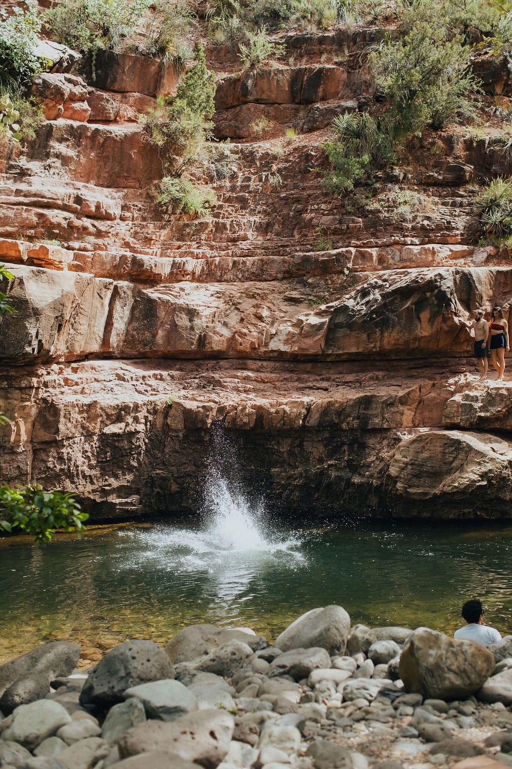 A splash disturbs the water at the base of a rocky cliff during one of their 36-Hour Adventures, with two people standing above and another sitting on stones by the edge of the pond, surrounded by greenery.