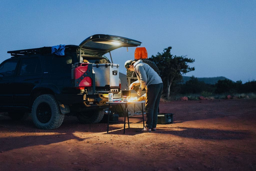 A person stands by an SUV with the trunk open, cooking at a portable table in a remote outdoor area at dusk—perfect for 36-Hour Adventures. Camping gear and a cooler are visible, capturing the spirit of spontaneous escapes and outdoor exploration.