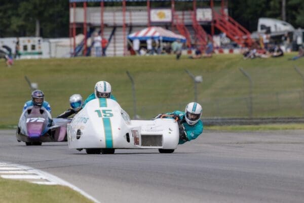 Three sidecar racing motorcycles speed around a racetrack, reminiscent of the excitement found at overland events and rallies. The front vehicle, number 15, has a passenger leaning out. Behind it, two more teams follow closely. Spectators and a red structure are visible in the background.