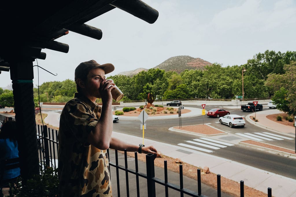 A person in a camouflage shirt and cap drinks from a cup while standing on a balcony, perhaps taking a break during their 36-Hour Adventures, overlooking a roundabout with cars and green trees in the background.
