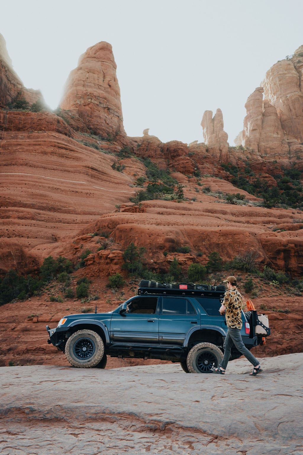 A person walks past a blue off-road SUV parked on rocky terrain, with towering red rock formations and cliffs in the background under a clear sky—the perfect setting for unforgettable 36-Hour Adventures.