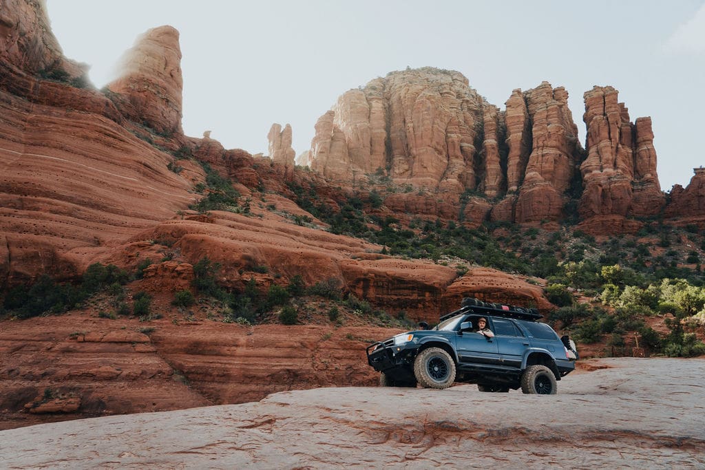 A black SUV with off-road tires drives on a rocky terrain with red sandstone cliffs and rock formations in the background under a bright sky, perfect for those seeking 36-Hour Adventures.