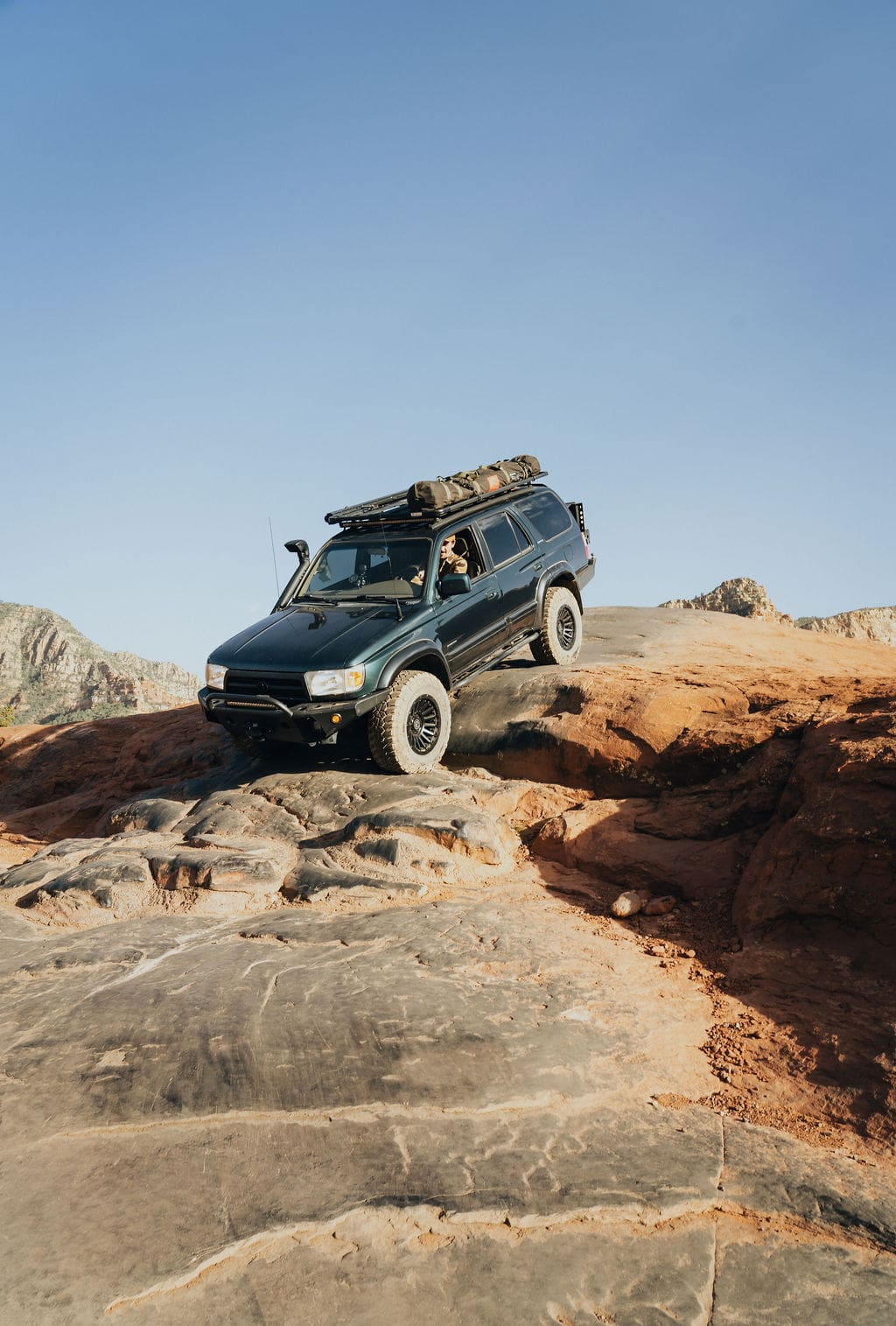 A black SUV with roof gear drives over rugged red rocks in a desert landscape under a clear blue sky, capturing the spirit of 36-Hour Adventures.