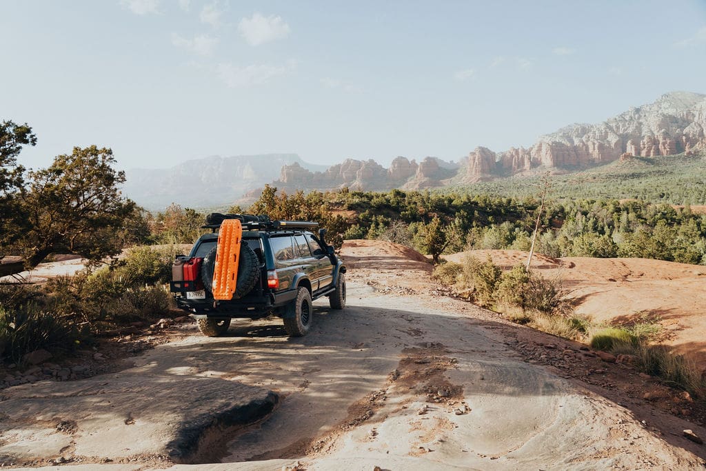A blue SUV with orange recovery boards on the roof drives on a rocky dirt road through a scenic desert landscape, embodying the spirit of 36-Hour Adventures amid striking red rock formations and patches of greenery in the background.