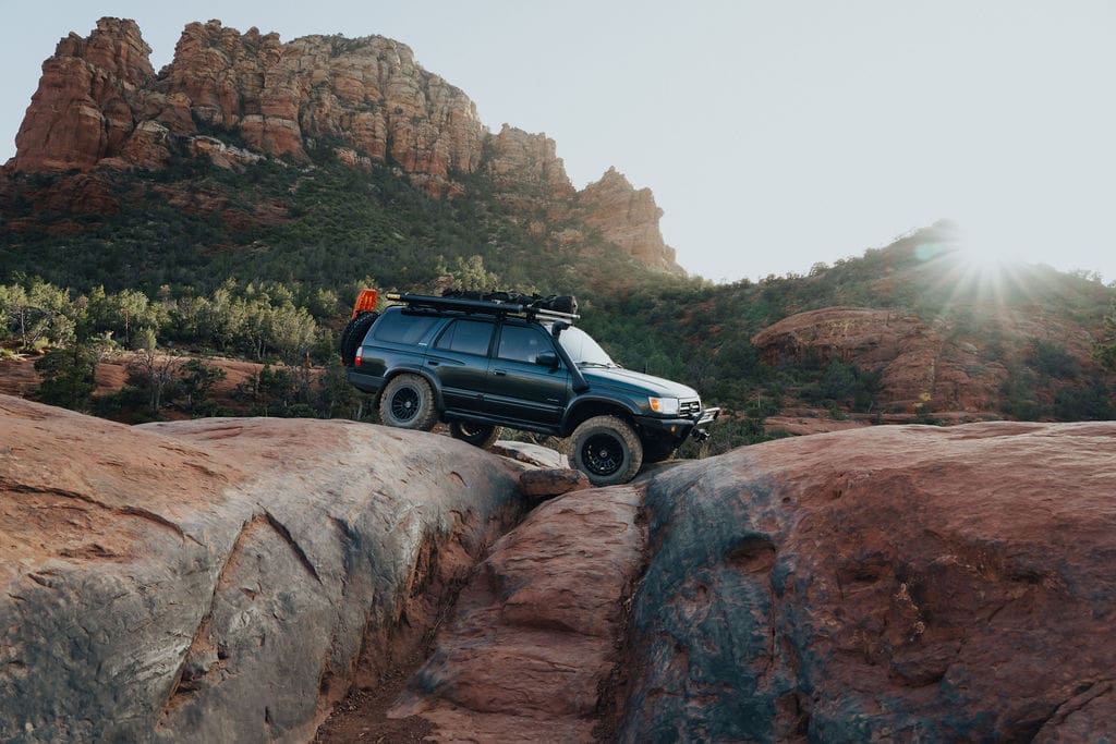 A dark SUV with off-road gear drives over large red rocks in a desert canyon landscape, perfect for 36-Hour Adventures amid rocky cliffs and the sun glowing in the background.
