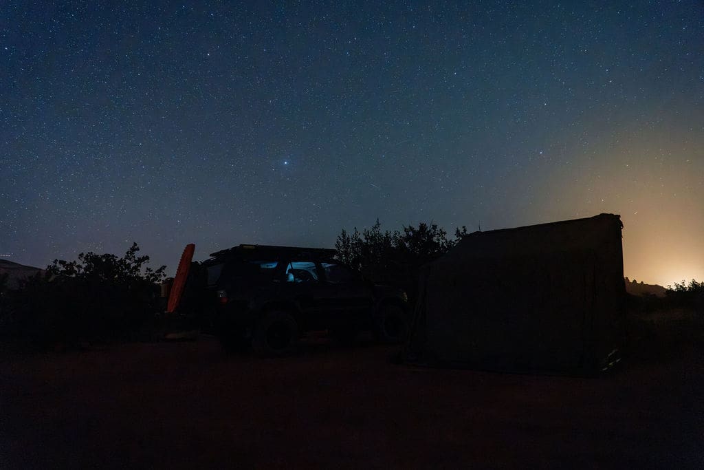 A car and a tent sit under a clear, starry night sky with a faint glow on the horizon, surrounded by silhouettes of trees and bushes—the perfect scene for unforgettable 36-Hour Adventures.