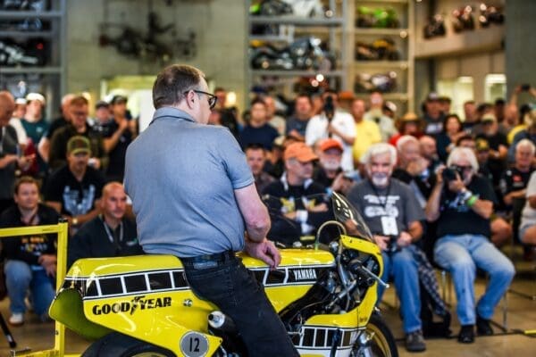 A man sits on a yellow Yamaha motorcycle in front of a seated crowd at an indoor overland event, with many people watching and taking photos—capturing the excitement typical of rallies and gatherings devoted to adventure riding.