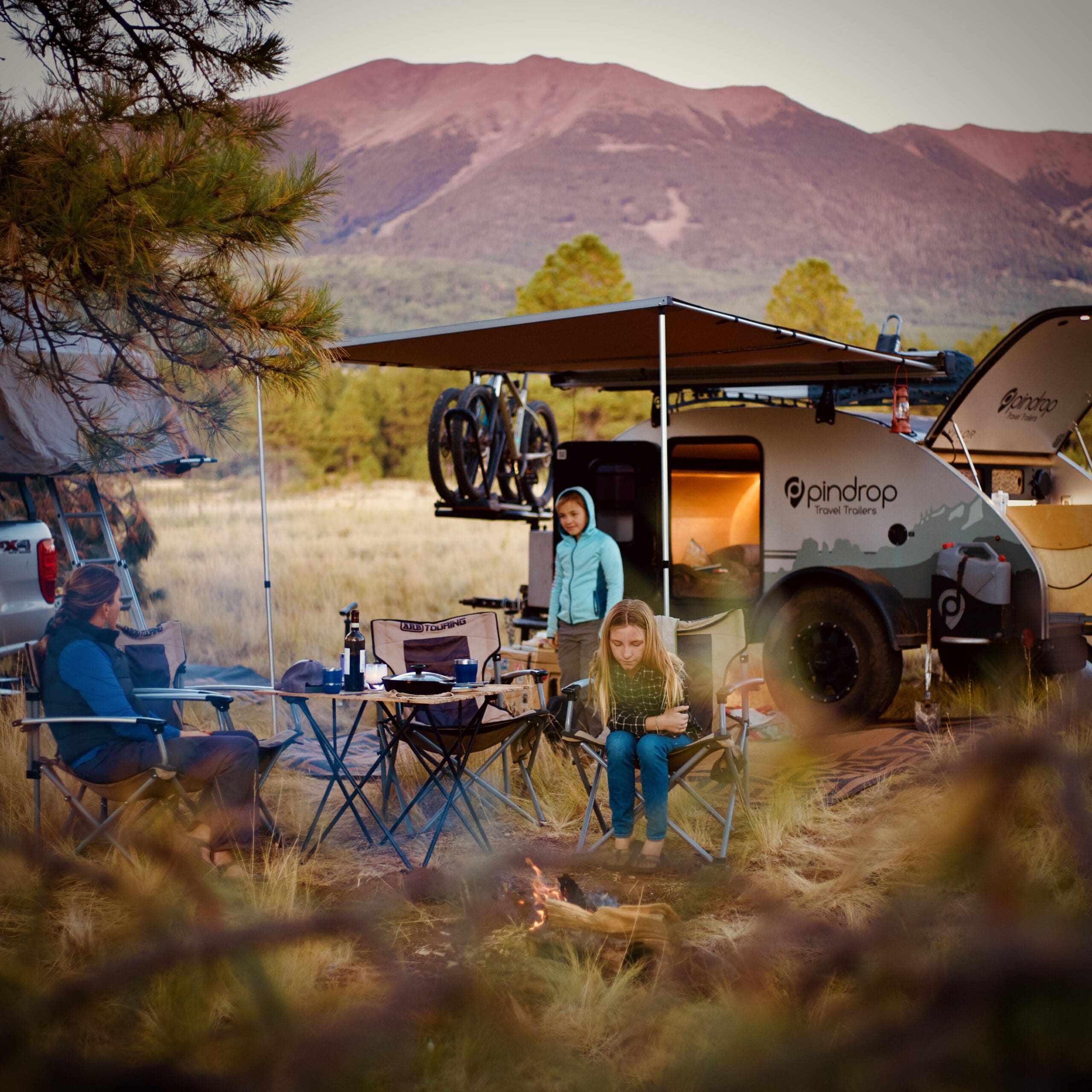 Three people sit and stand around a campfire near a small travel trailer, with bikes mounted on top. They are surrounded by grass, trees, and distant mountains during sunset—a classic scene often found at overland events and rallies. Camping chairs and gear are scattered nearby.