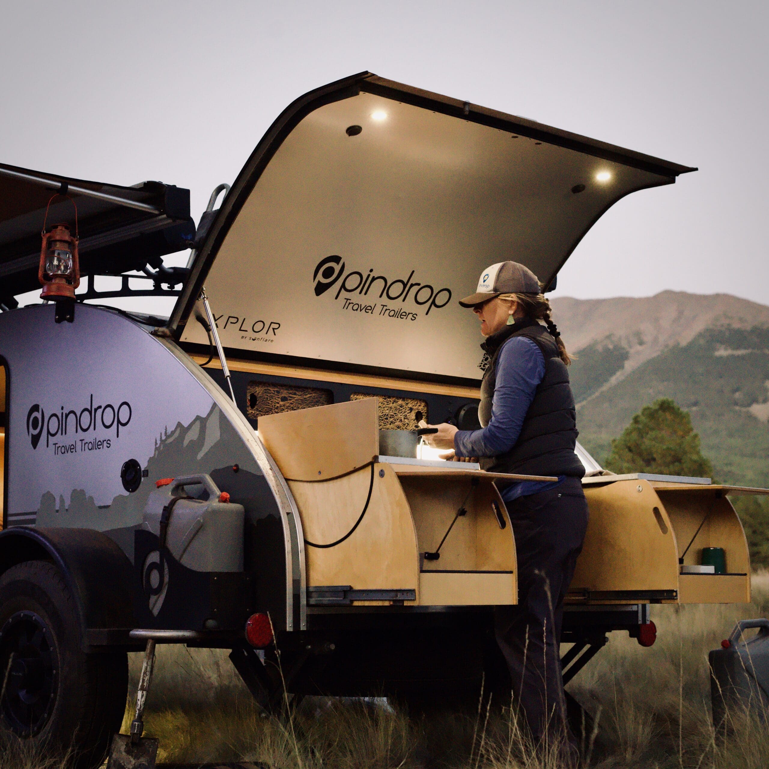 A person stands at the open back of a teardrop trailer, preparing food on a pull-out kitchen, surrounded by nature with hills and trees in the background—a perfect scene often found at overland events and rallies.