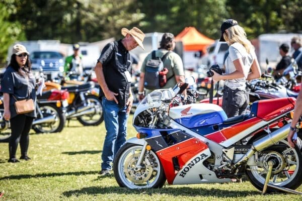 People gather outdoors at a motorcycle show, admiring various bikes. A Honda motorcycle with red, white, and blue colors is in the foreground, and several individuals are standing around, observing and taking photos. The lively atmosphere is reminiscent of overland events and rallies, where enthusiasts come together to celebrate their shared passion for adventure on two wheels.