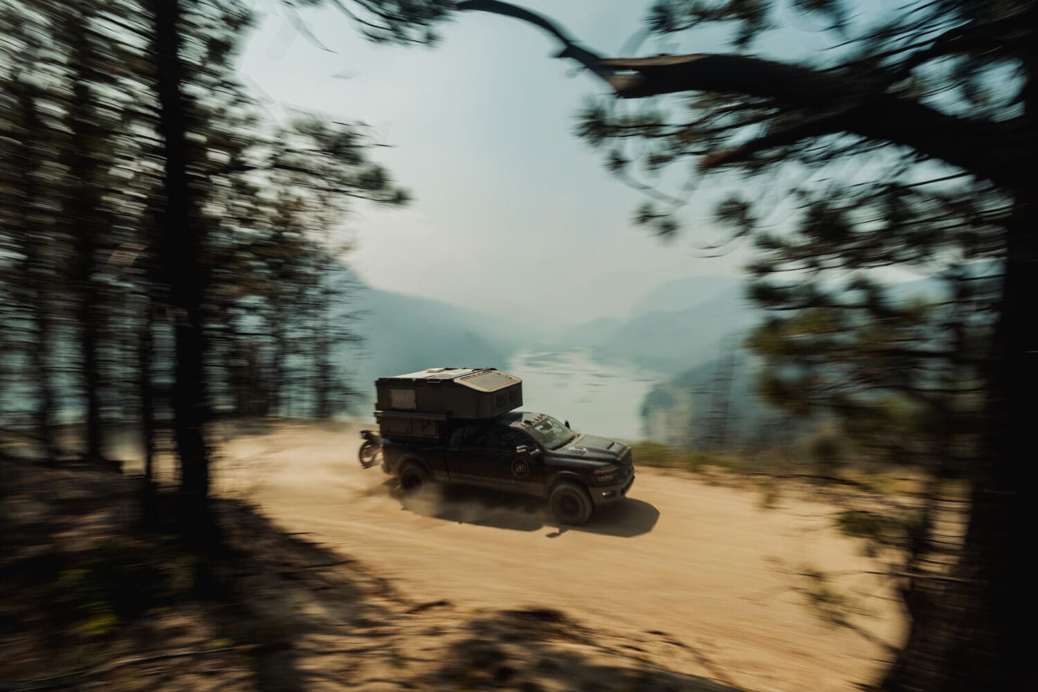 A black off-road truck with a Limited Edition Scout Camper speeds along a dirt road through a pine forest, with mountains and a river visible in the misty Sitka background.