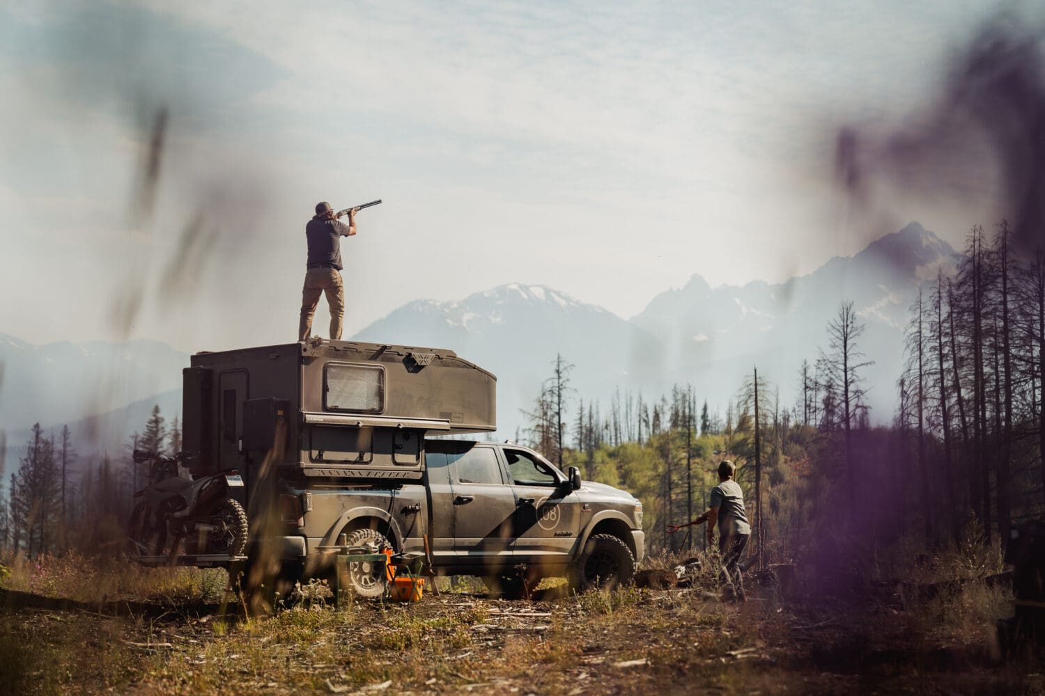 A person stands on top of a rugged Scout Camper truck aiming binoculars, while another stands nearby in a forest clearing with mountains in the background. The scene is adventurous and outdoorsy.
