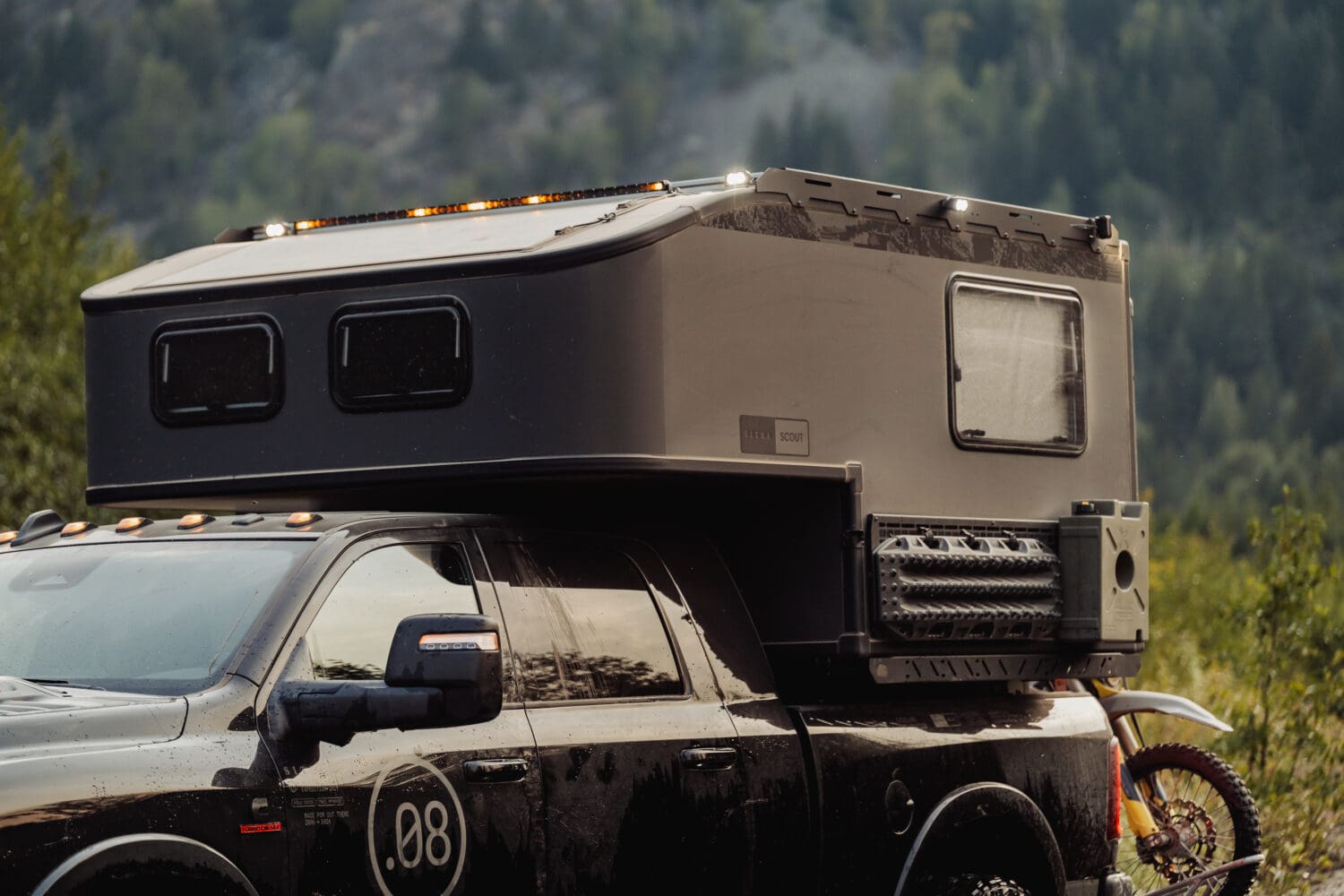 A black truck with a Limited Edition Scout Camper on its bed is parked outdoors. The camper boasts several windows, mounted gear, and is surrounded by greenery and mountains in the background.